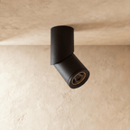 A black adjustable cylindrical downlight is angled from a plaster ceiling corner, its lens glowing warm, a Clérachic fixture close up.