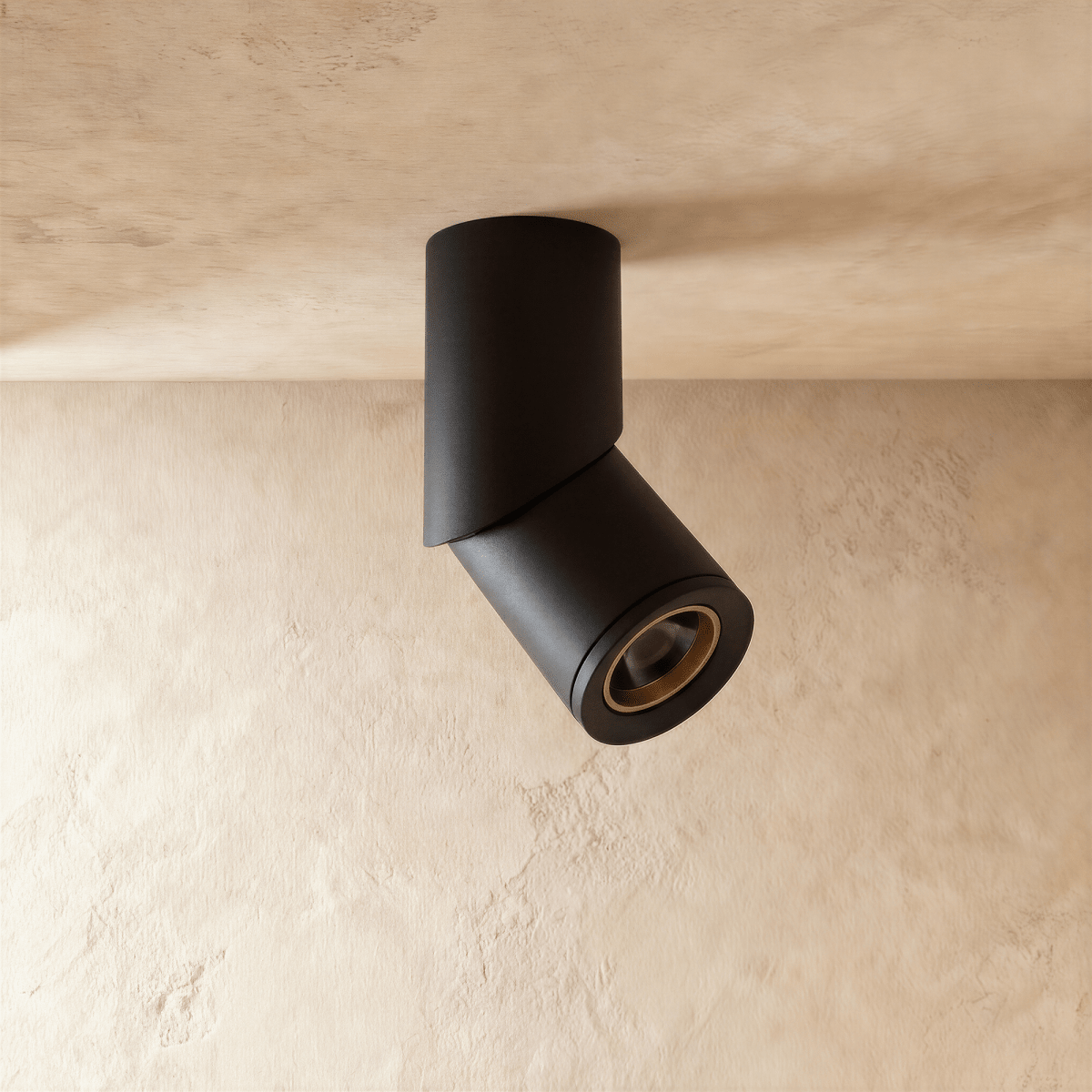 A black adjustable cylindrical downlight is angled from a plaster ceiling corner, its lens glowing warm, a Clérachic fixture close up.