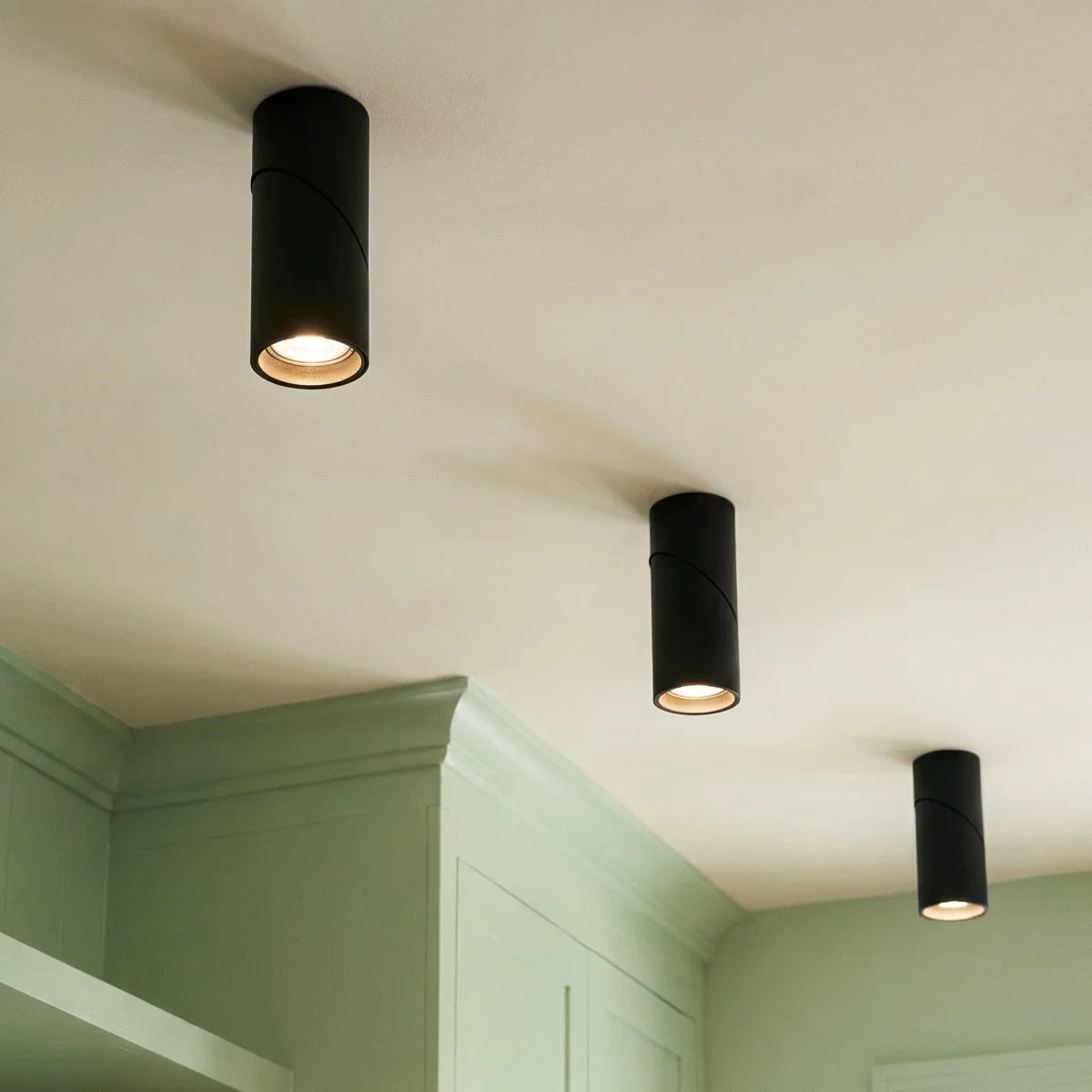 Three black adjustable downlights glow above sage green kitchen cabinetry, their lenses angled downward, Clérachic fixtures close up.