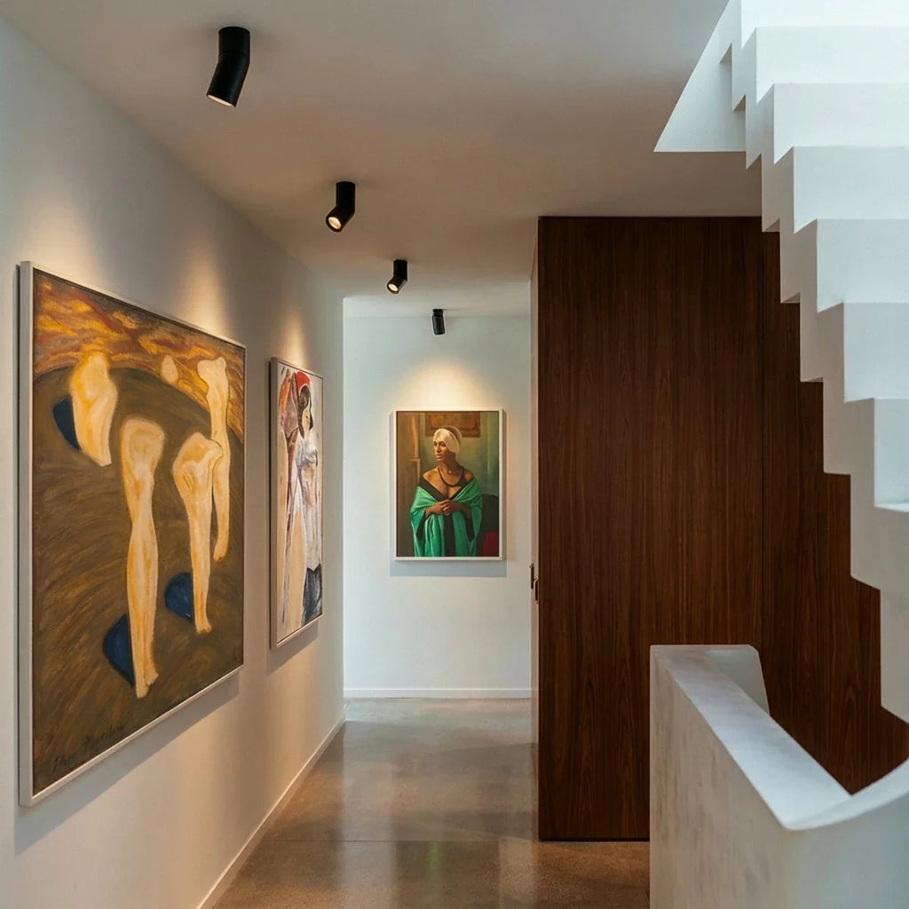 Black adjustable downlights line a gallery hallway ceiling beside a staircase and framed paintings, these Clérachic fixtures angled.
