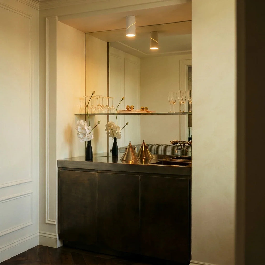 A white adjustable downlight lights a mirrored bar nook with glassware and a dark metal counter, this Clérachic fixture overhead.