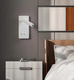 Clérachic reading light in white mounted beside a bed, books stacked on the nightstand below.