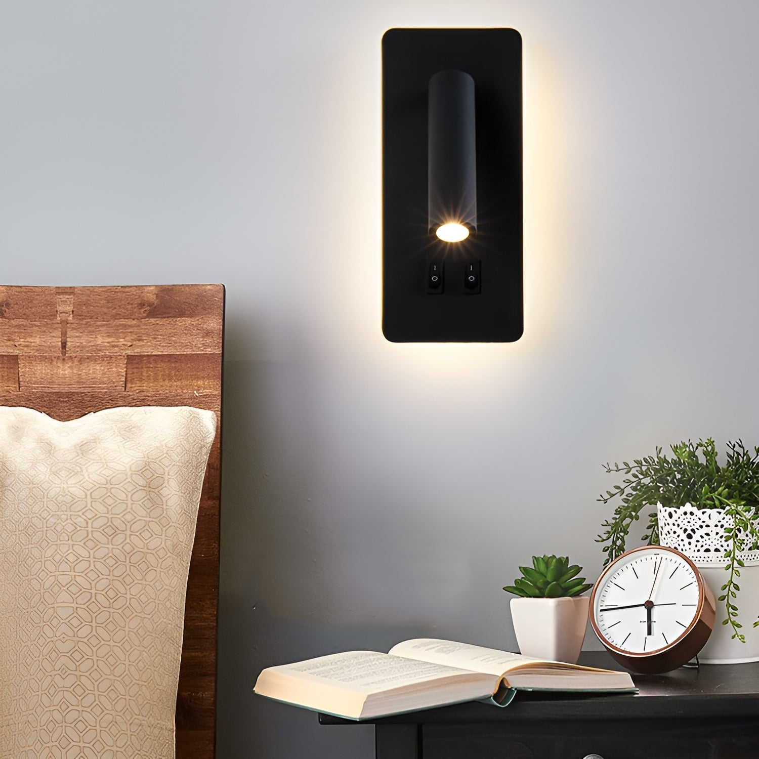 The black Clérachic reading light lit above a bedside table with a clock and small plants.