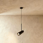 Clérachic black cylinder pendant on a thin cord with an angled spot head, lit warm against a limewash wall.