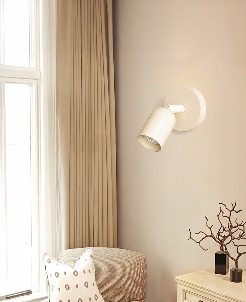 A cream-white Clérachic wall light angled on a beige wall beside a curtain, with an armchair and a branch in a vase below.