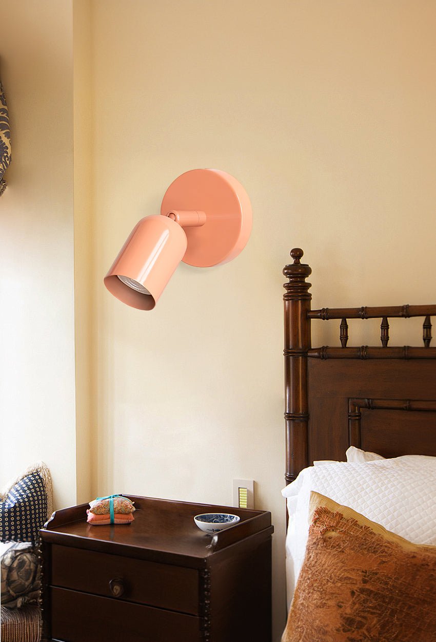 Above a dark wooden bedside chest and a carved bed, a pink Clérachic wall light is tilted down on a warm cream wall.