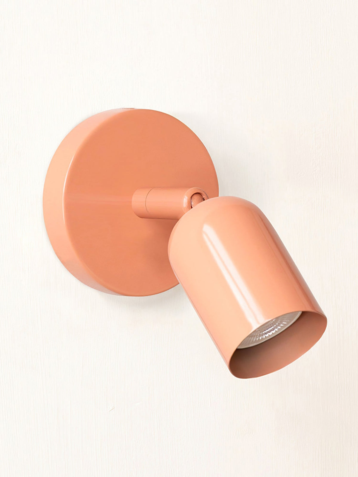 A pink Clérachic wall light with a round backplate and an adjustable cylindrical head, the bulb visible inside, on cream.