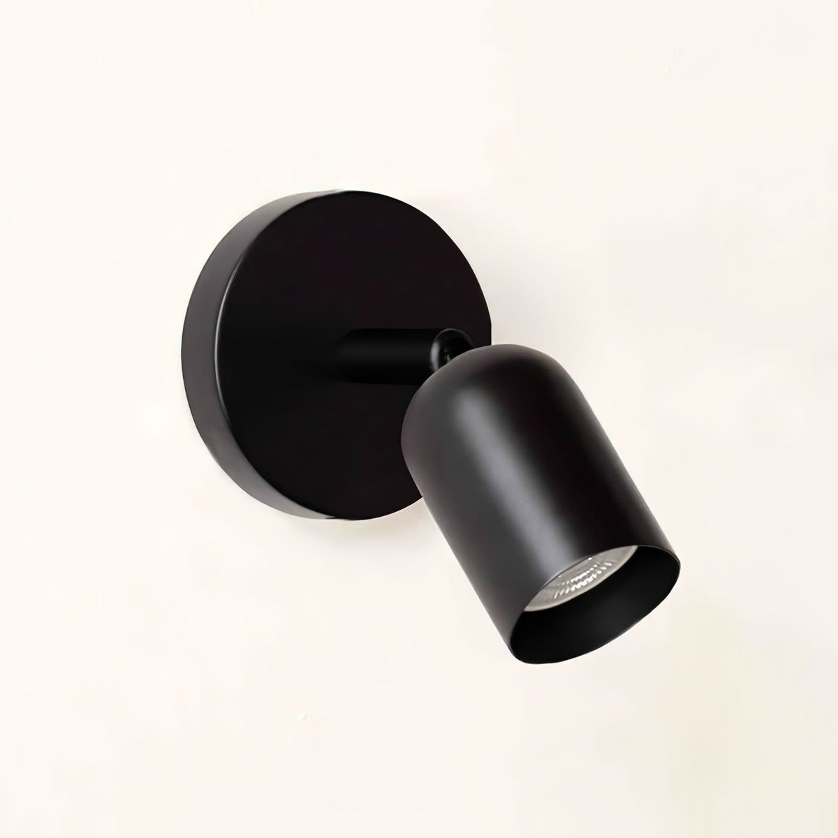 Matte black Clérachic wall light with a round backplate and a tilted cylindrical head, shown on a pale cream wall.