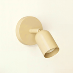 Soft cream-beige Clérachic wall light with a round plate and a short tilting head, seen against a warm cream wall.