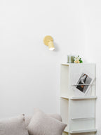 Small in the frame, a butter-yellow Clérachic wall light glows above a white shelf unit with a candle jar and flowers.