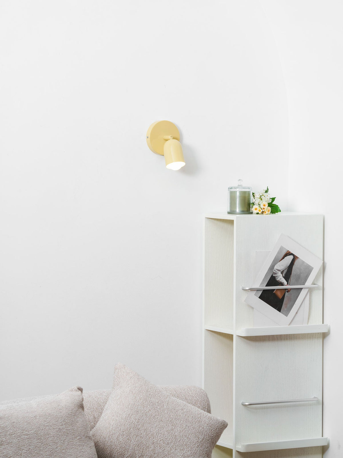 Small in the frame, a butter-yellow Clérachic wall light glows above a white shelf unit with a candle jar and flowers.