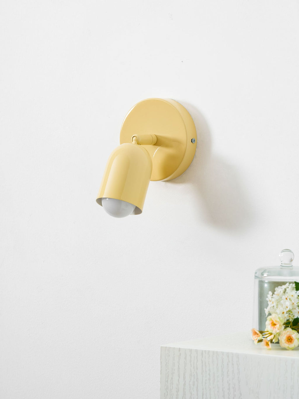 A butter-yellow Clérachic wall light with its head tilted down, lit, beside a glass cloche of flowers on a white ledge.