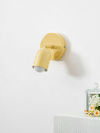 A butter-yellow Clérachic wall light with its head tilted down, lit, beside a glass cloche of flowers on a white ledge.