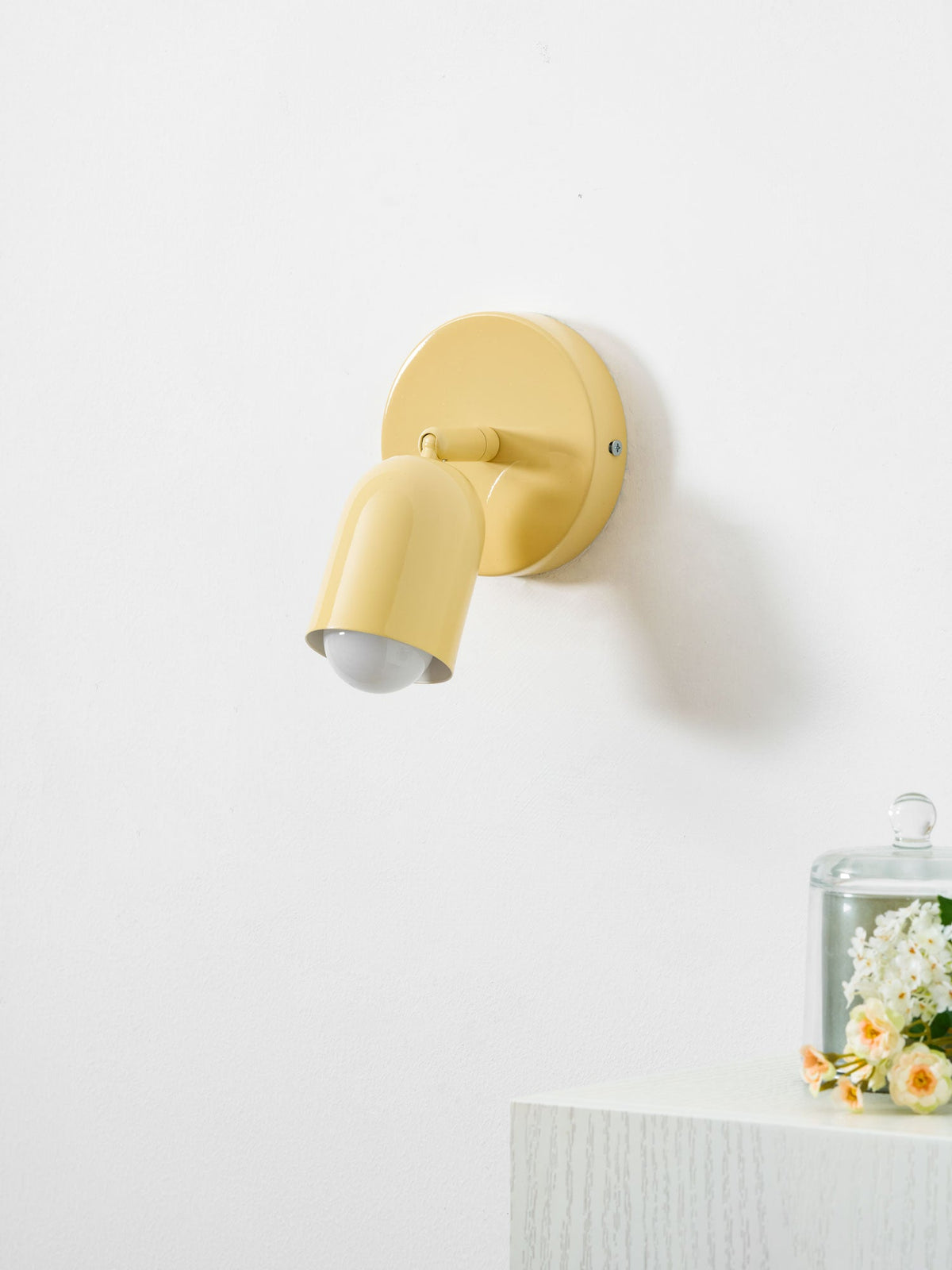 A butter-yellow Clérachic wall light with its head tilted down, lit, beside a glass cloche of flowers on a white ledge.