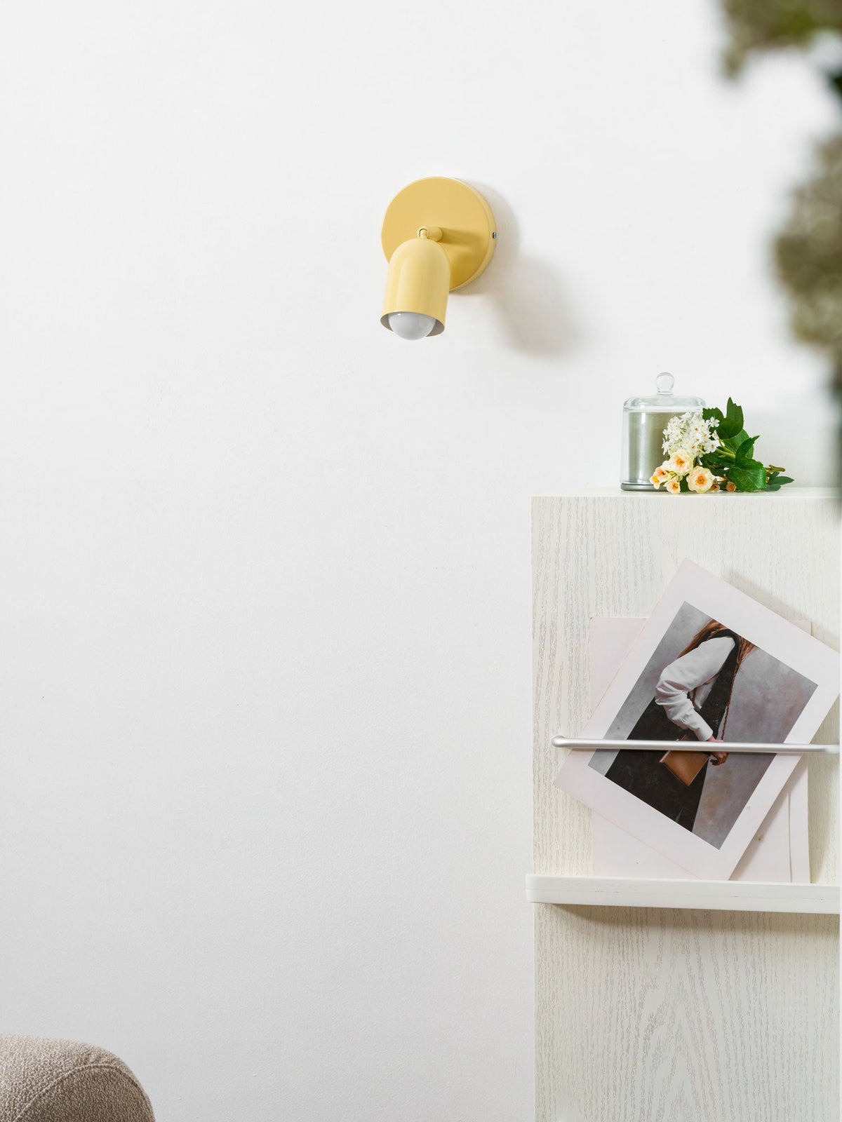 A pale yellow Clérachic wall light sits high on a white wall above a white shelf holding a candle, flowers and a print.