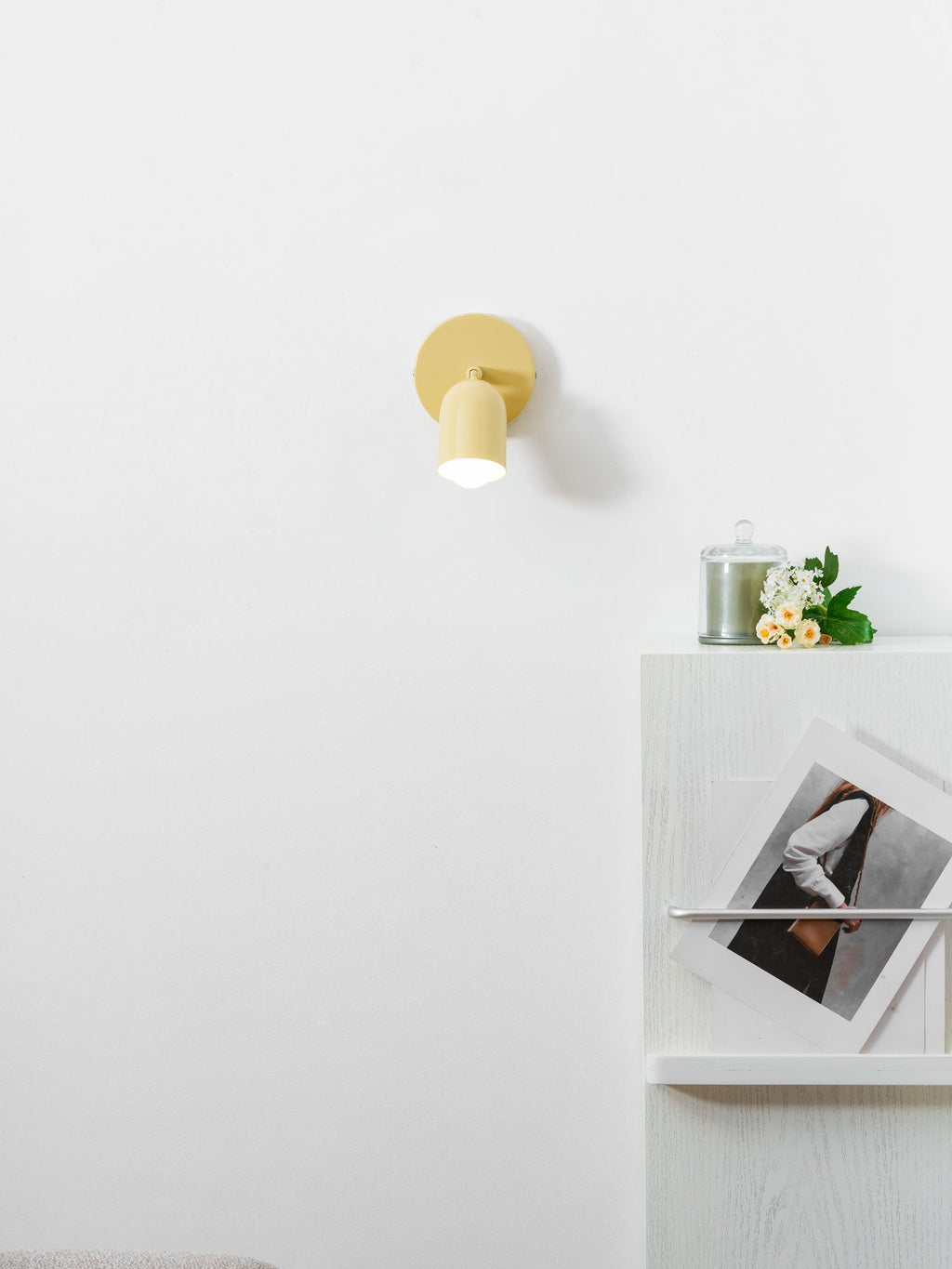 Lit yellow Clérachic wall light above a white cabinet with a candle, a bunch of flowers and a framed print leaning on it.