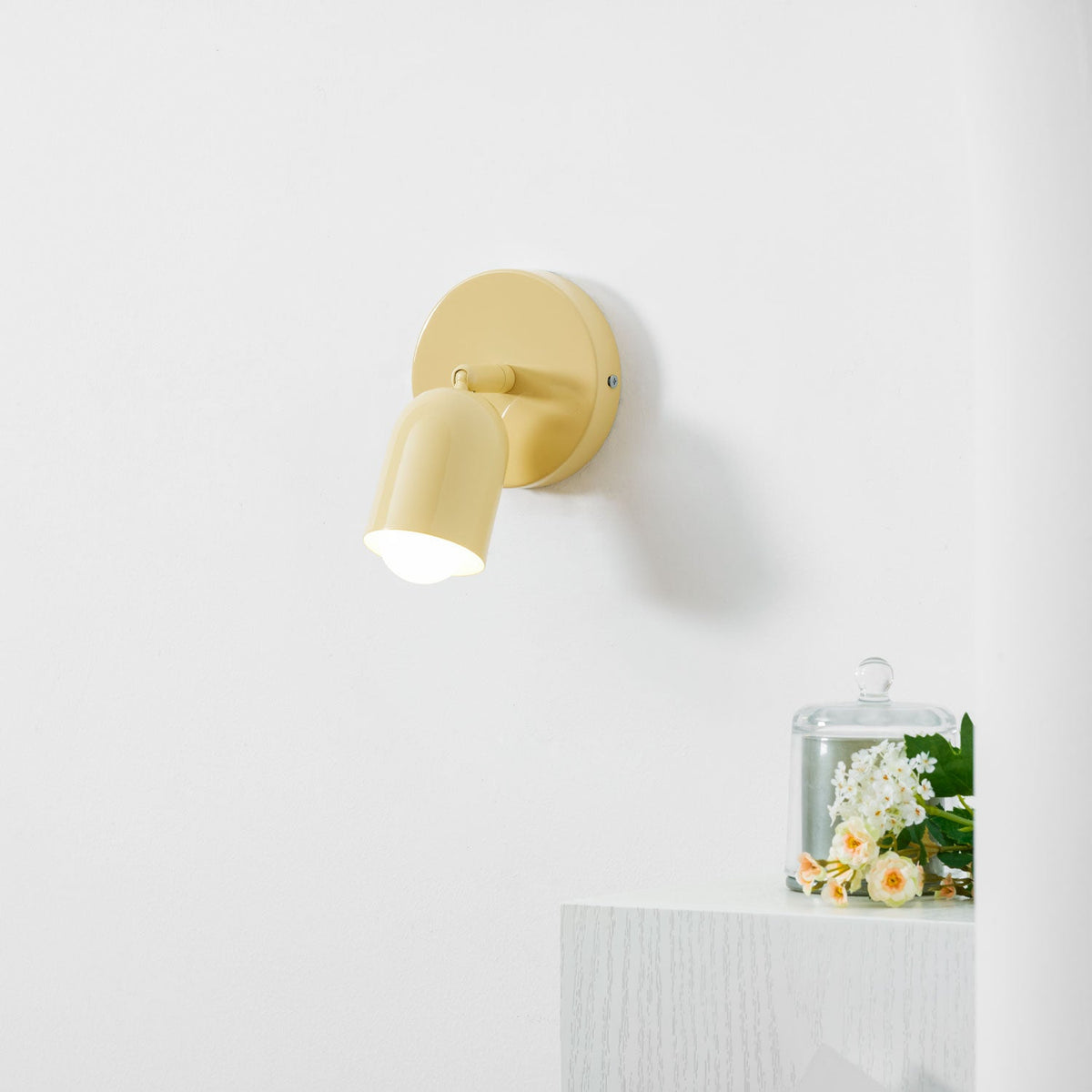 A yellow Clérachic wall light with its head swung down and lit, on a white wall above a ledge with a glass cloche.