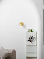 Against a white arched wall, a small yellow Clérachic wall light shines above a shelf with flowers and a leaning print.