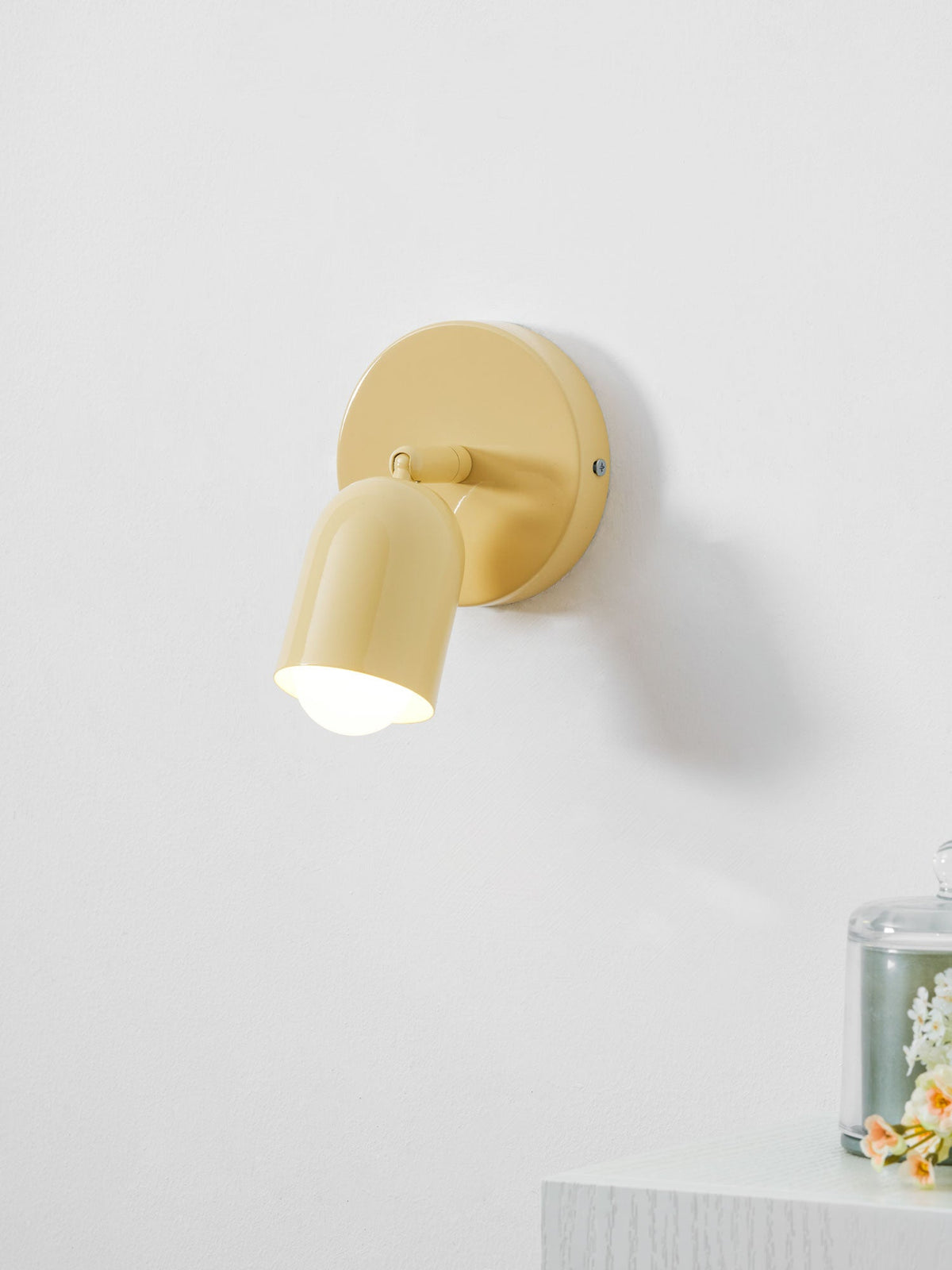 Close view of a butter-yellow Clérachic wall light, its tilted head glowing, above a white ledge with flowers under glass.