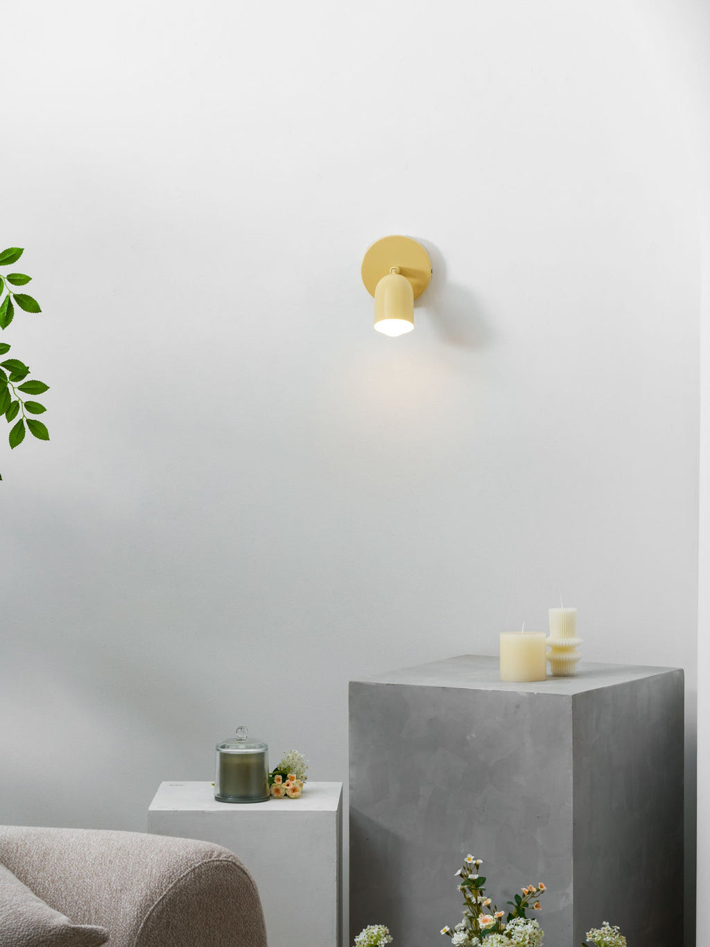 Lit yellow Clérachic wall light on a white wall above a grey stone block, with candles and a green plant to the left.