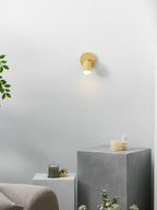 Lit yellow Clérachic wall light on a white wall above a grey stone block, with candles and a green plant to the left.