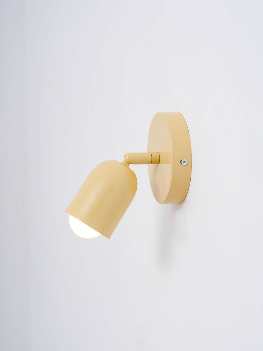 A butter-yellow Clérachic wall light with a lit, tilted head seen from the left on a pale grey wall.