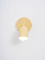 Front view of a butter-yellow Clérachic wall light with a lit bulb inside the cylindrical head, on a pale grey wall.