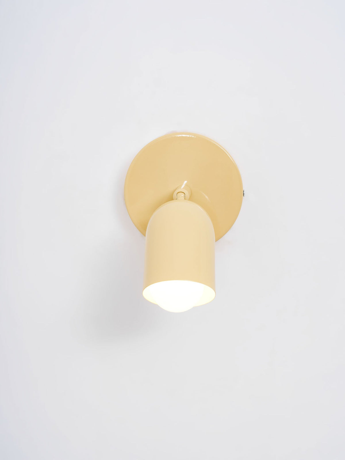 Front view of a butter-yellow Clérachic wall light with a lit bulb inside the cylindrical head, on a pale grey wall.