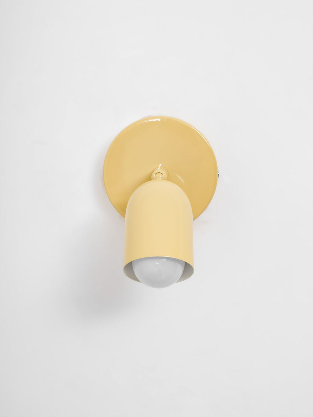 Butter-yellow Clérachic wall light with its head aimed straight down, the round bulb visible in the opening, on a pale wall.