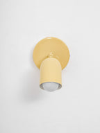 Butter-yellow Clérachic wall light with its head aimed straight down, the round bulb visible in the opening, on a pale wall.