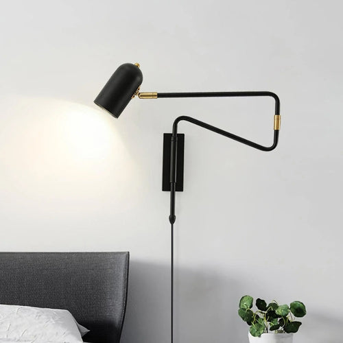 Clérachic adjustable wall light in black with gold collars, throwing a warm pool of light on the wall above a grey upholstered headboard, with a round-leafed plant in a white pot below.