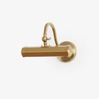 Gold Clérachic picture light with a hexagonal bar shade and a curved swan-neck arm, on a round wall plate, against a plain white wall.