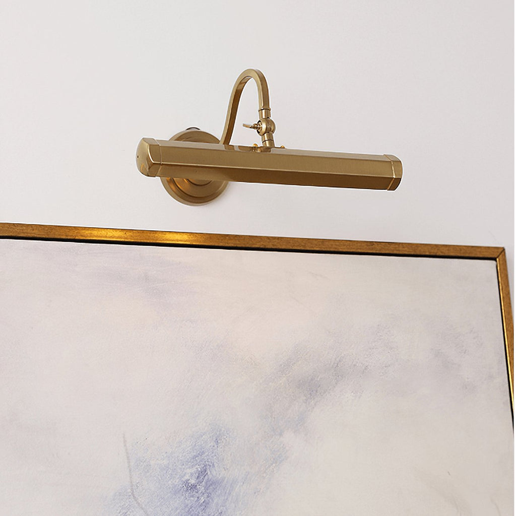 A gold Clérachic wall light sits just above a framed pale abstract painting, its bar shade held on a curved arm.