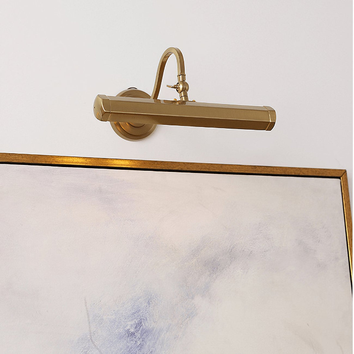 A gold Clérachic wall light sits just above a framed pale abstract painting, its bar shade held on a curved arm.