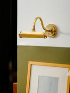 Beside a dark doorway and an olive-green wall, this gold Clérachic wall light sits above a framed print in a gold frame.