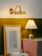 Warm light from a gold Clérachic wall light falls on an olive wall, with a framed print and a pleated-shade table lamp below.