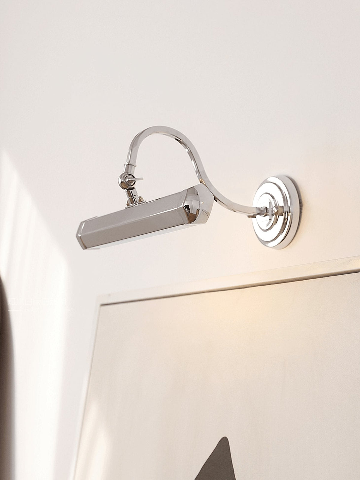 Chrome wall light from Clérachic tilted toward a pale artwork on a cream wall, with a warm glow from the shade.