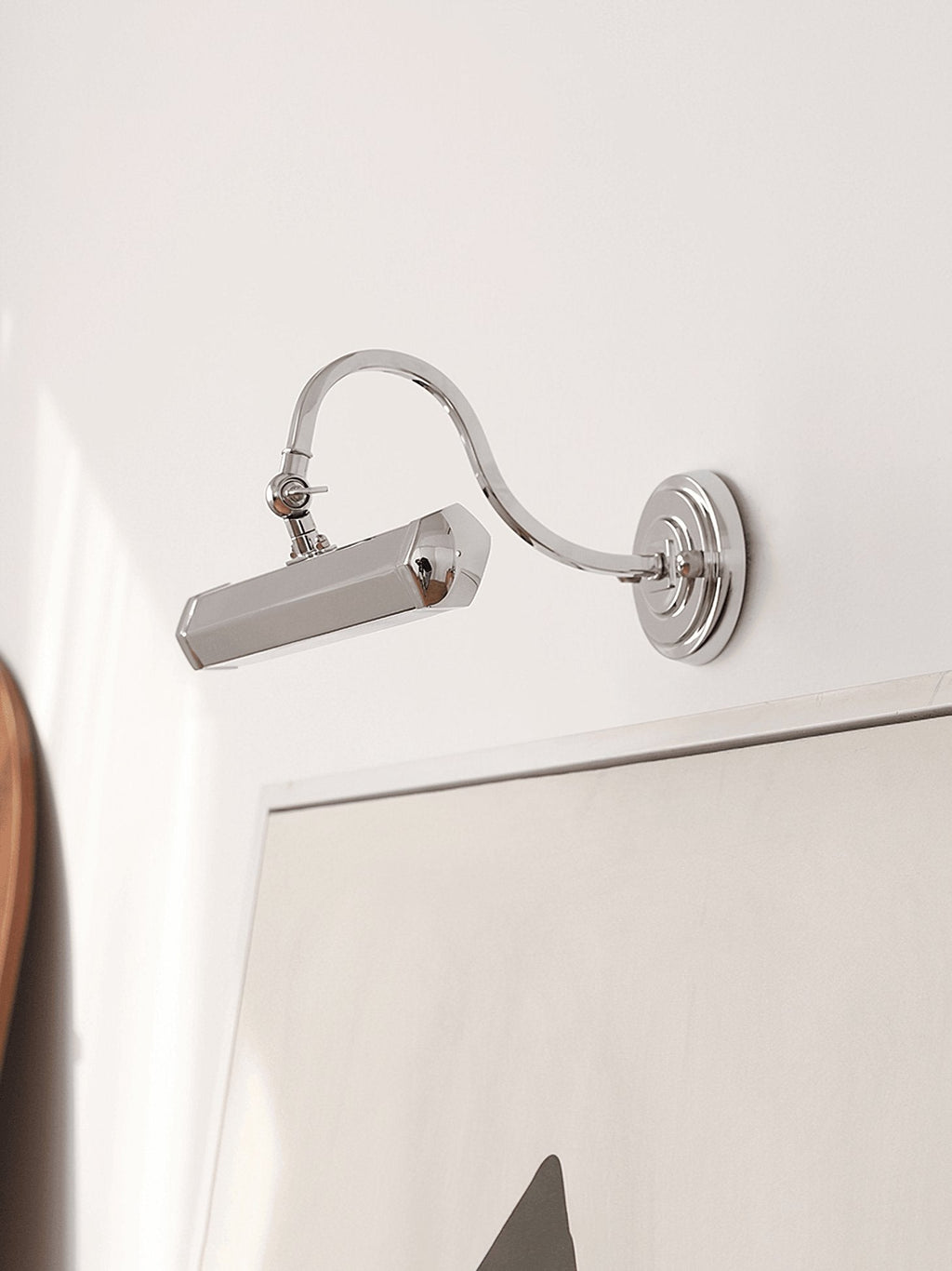 Chrome Clérachic wall light with a curved arm, tilted down beside a white frame, on a pale wall.
