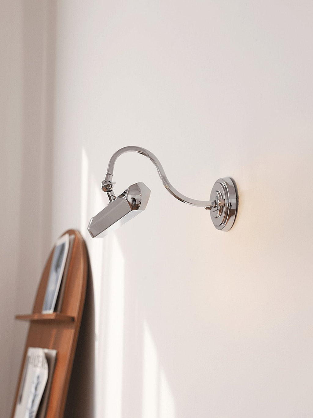 On a pale wall beside a wooden shelf, this chrome Clérachic wall light is angled downward on its curved arm.