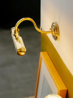 Gold Clérachic wall light angled steeply toward a gold picture frame, with its shade catching warm light.
