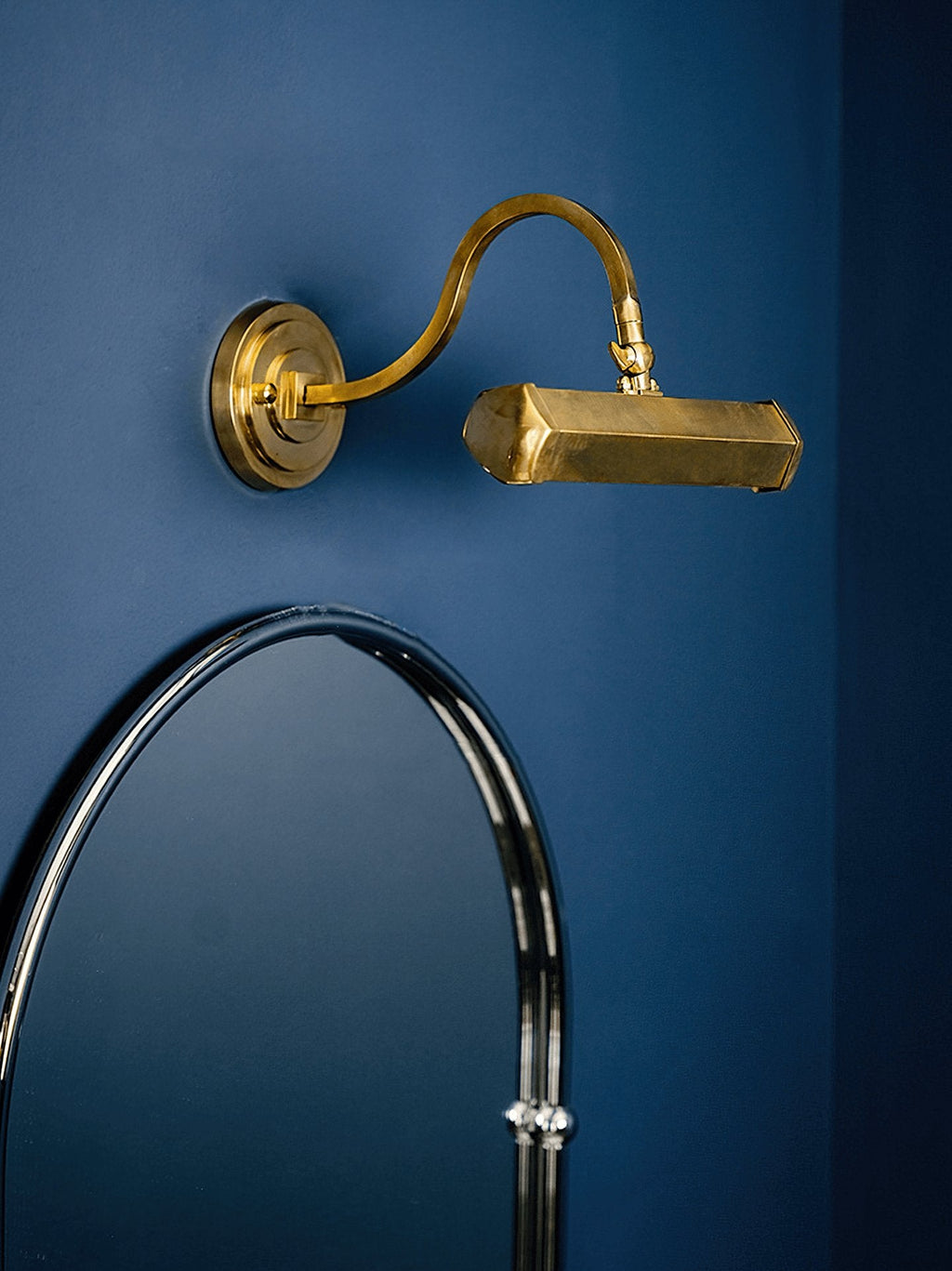 Gold Clérachic wall light on a deep blue wall above an arched mirror, with its curved arm reaching out to the shade.