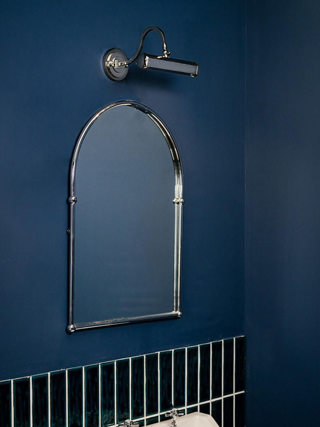 A chrome wall light from Clérachic sits above an arched chrome mirror on a navy wall, with green tiles below.