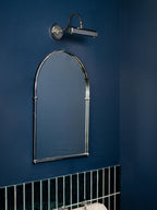 A chrome wall light from Clérachic sits above an arched chrome mirror on a navy wall, with green tiles below.