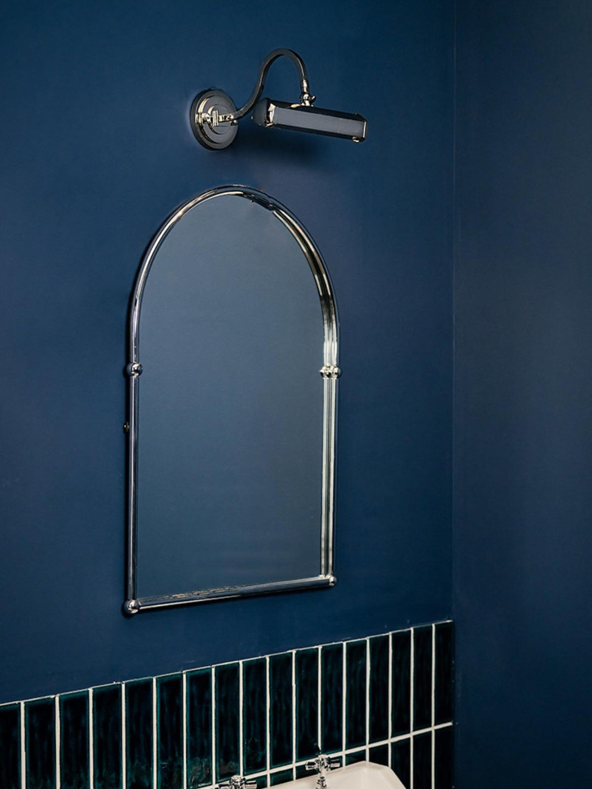 A chrome wall light from Clérachic sits above an arched chrome mirror on a navy wall, with green tiles below.
