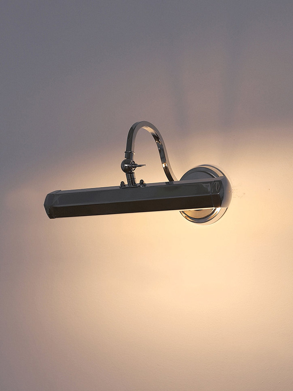 A chrome Clérachic wall light in a dim room, casting a wide pool of warm light on the wall around it.
