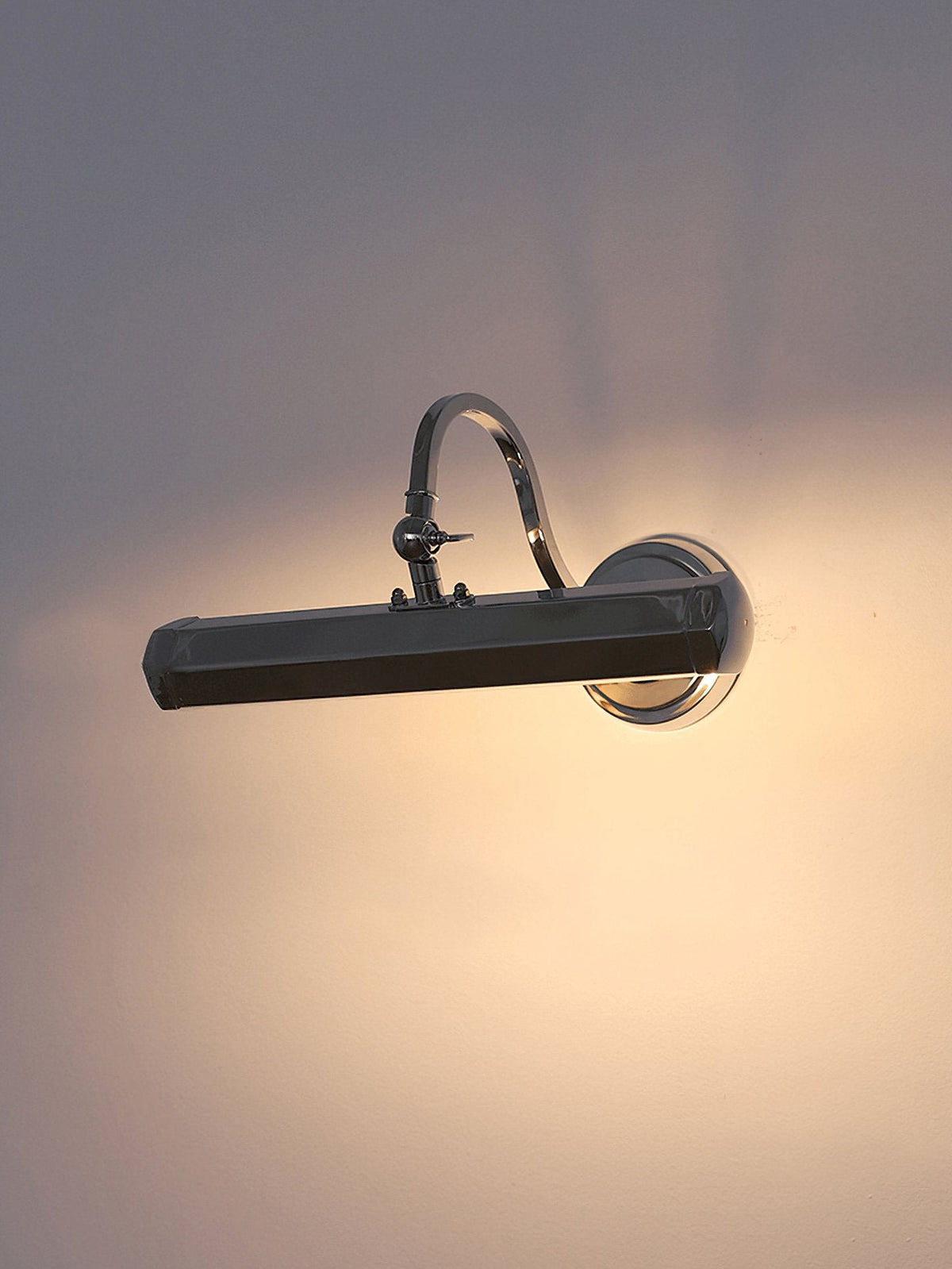 A chrome Clérachic wall light in a dim room, casting a wide pool of warm light on the wall around it.