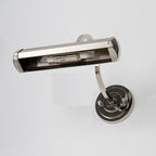 Seen from slightly below, a chrome Clérachic wall light with its shade open to show the tube bulb inside, on a pale wall.