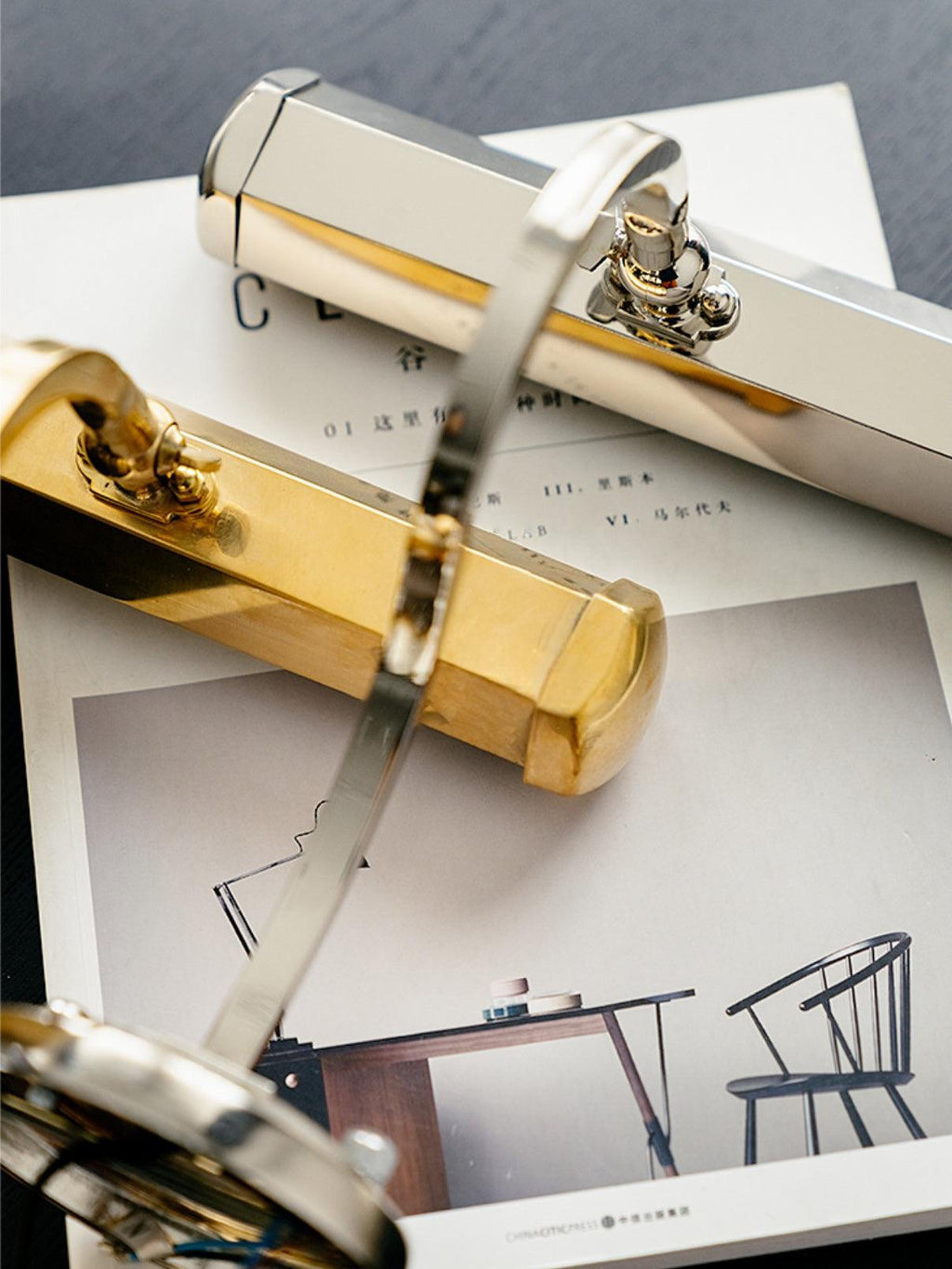Two Clérachic wall lights, one gold and one chrome, lying on a printed magazine to show the two finishes side by side.