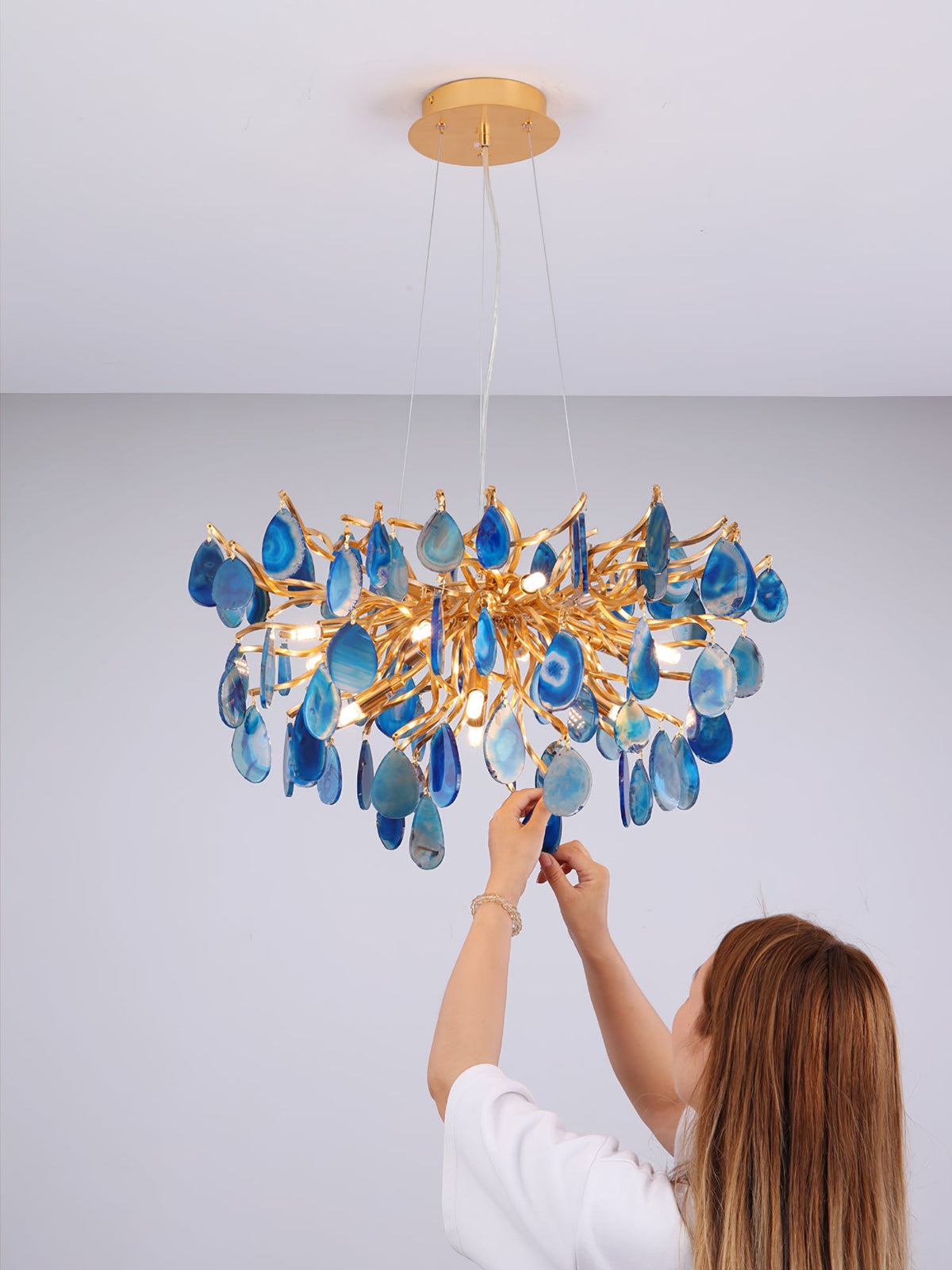 Blue agate Clérachic chandelier on a lilac wall, with a woman reaching up to adjust one of the stone slices.