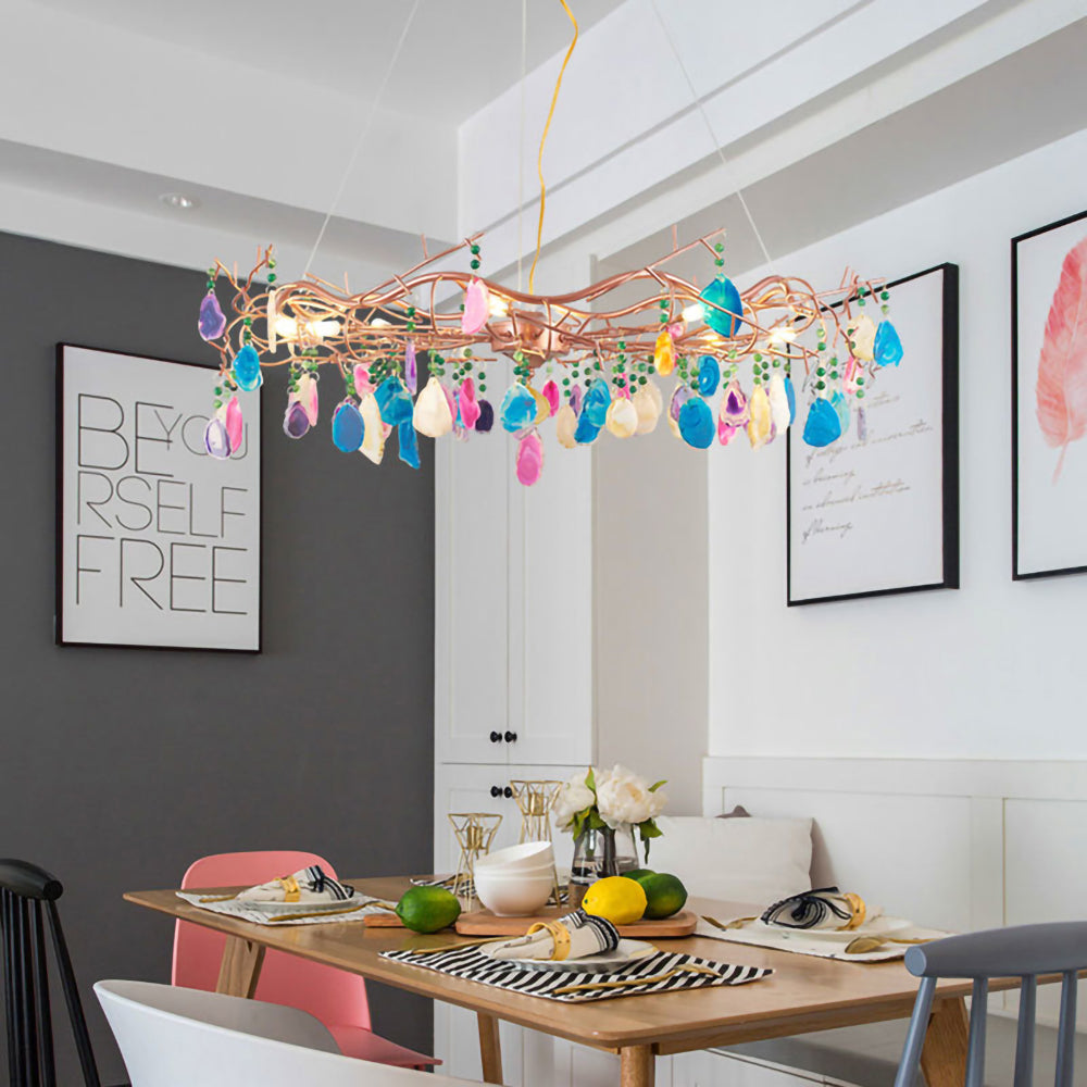 This Clérachic agate chandelier hangs over a wooden dining table set with fruit, in a room with a dark grey wall and a framed poster.